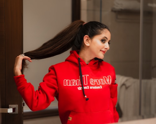 A person is standing indoors, looking at their reflection in a mirror. They are wearing a bright red hooded sweatshirt and have long brown hair styled in a high ponytail. The hand is reaching back to hold the ponytail. The background appears to be a bathroom, with towels visible.