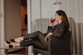 A woman sits comfortably on a woven chair, dressed in a stylish dark pantsuit. She is sipping a red drink from a clear glass, with sunglasses resting on her head. Her posture is relaxed, with legs propped up on a small round table. The background features light-colored curtains and a glimpse into a warmly lit room.