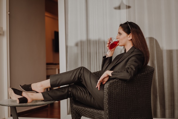 A woman sits comfortably on a woven chair, dressed in a stylish dark pantsuit. She is sipping a red drink from a clear glass, with sunglasses resting on her head. Her posture is relaxed, with legs propped up on a small round table. The background features light-colored curtains and a glimpse into a warmly lit room.