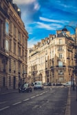 A picturesque urban street lined with classic Parisian architectural buildings under a vivid blue sky. The street is paved with cobblestones and features several vehicles, including cars and vans, driving along. Elegant street lamps and traffic signs are visible, adding to the atmosphere of typical city life.