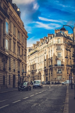 A picturesque urban street lined with classic Parisian architectural buildings under a vivid blue sky. The street is paved with cobblestones and features several vehicles, including cars and vans, driving along. Elegant street lamps and traffic signs are visible, adding to the atmosphere of typical city life.