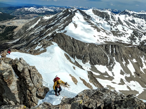 Two climbers ascend a snowy, rocky mountain ridge with snow-covered peaks and valleys in the background. The climbers are wearing appropriate mountain climbing gear including jackets, helmets, and backpacks. The scene is set in a mountainous, high-altitude environment under a partly cloudy sky.