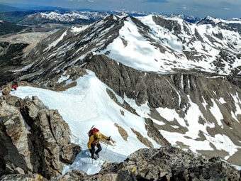 Two climbers ascend a snowy, rocky mountain ridge with snow-covered peaks and valleys in the background. The climbers are wearing appropriate mountain climbing gear including jackets, helmets, and backpacks. The scene is set in a mountainous, high-altitude environment under a partly cloudy sky.