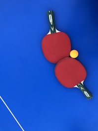 Two red ping pong paddles are placed on a vibrant blue surface, with a bright yellow ping pong ball positioned near them. The handles of the paddles have a green and black design with a white label featuring the brand 'DUNLOP'. A thin white line is visible in the lower-left corner, likely part of a table tennis table.