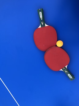 Two red ping pong paddles are placed on a vibrant blue surface, with a bright yellow ping pong ball positioned near them. The handles of the paddles have a green and black design with a white label featuring the brand 'DUNLOP'. A thin white line is visible in the lower-left corner, likely part of a table tennis table.