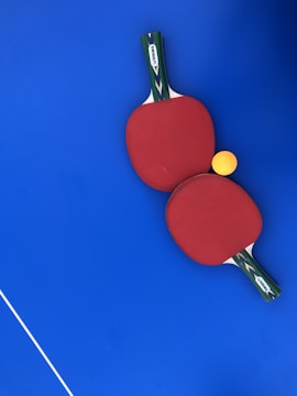 Two red ping pong paddles are placed on a vibrant blue surface, with a bright yellow ping pong ball positioned near them. The handles of the paddles have a green and black design with a white label featuring the brand 'DUNLOP'. A thin white line is visible in the lower-left corner, likely part of a table tennis table.