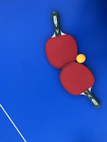 Two red ping pong paddles are placed on a vibrant blue surface, with a bright yellow ping pong ball positioned near them. The handles of the paddles have a green and black design with a white label featuring the brand 'DUNLOP'. A thin white line is visible in the lower-left corner, likely part of a table tennis table.