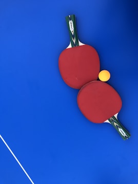 Two red ping pong paddles are placed on a vibrant blue surface, with a bright yellow ping pong ball positioned near them. The handles of the paddles have a green and black design with a white label featuring the brand 'DUNLOP'. A thin white line is visible in the lower-left corner, likely part of a table tennis table.