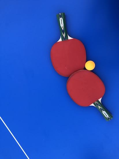 Two red ping pong paddles are placed on a vibrant blue surface, with a bright yellow ping pong ball positioned near them. The handles of the paddles have a green and black design with a white label featuring the brand 'DUNLOP'. A thin white line is visible in the lower-left corner, likely part of a table tennis table.