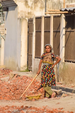 A woman stands amidst broken bricks and rubble, holding a tool. She is wearing a colorful dress with polka dots and a headscarf, leaning slightly on the tool with an expression of determination. The setting appears to be a street, bordered by a row of buildings with aged and peeling walls.