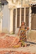 A woman stands amidst broken bricks and rubble, holding a tool. She is wearing a colorful dress with polka dots and a headscarf, leaning slightly on the tool with an expression of determination. The setting appears to be a street, bordered by a row of buildings with aged and peeling walls.