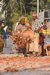 Several people are engaged in manual labor, handling bricks and rubble on a dusty road. One person is in mid-motion, dropping a basket of bricks.