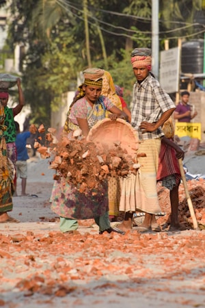 Several people are engaged in manual labor, handling bricks and rubble on a dusty road. One person is in mid-motion, dropping a basket of bricks.