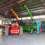 A warehouse setting contains several heavy machinery vehicles including a red vehicle with large tires, a green telescopic handler with an extended boom, and an orange excavator with a visible grabber attachment. The warehouse has a high ceiling with metal beams and ample light streaming through windows.