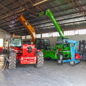 A warehouse setting contains several heavy machinery vehicles including a red vehicle with large tires, a green telescopic handler with an extended boom, and an orange excavator with a visible grabber attachment. The warehouse has a high ceiling with metal beams and ample light streaming through windows.
