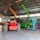 A warehouse setting contains several heavy machinery vehicles including a red vehicle with large tires, a green telescopic handler with an extended boom, and an orange excavator with a visible grabber attachment. The warehouse has a high ceiling with metal beams and ample light streaming through windows.