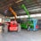 A warehouse setting contains several heavy machinery vehicles including a red vehicle with large tires, a green telescopic handler with an extended boom, and an orange excavator with a visible grabber attachment. The warehouse has a high ceiling with metal beams and ample light streaming through windows.