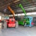 A warehouse setting contains several heavy machinery vehicles including a red vehicle with large tires, a green telescopic handler with an extended boom, and an orange excavator with a visible grabber attachment. The warehouse has a high ceiling with metal beams and ample light streaming through windows.