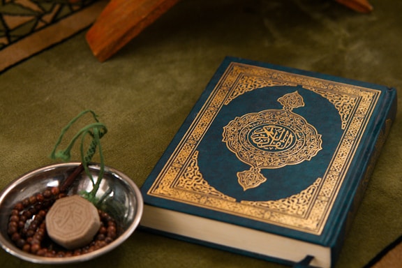 A Quran with an ornate cover is placed on a green fabric. Beside it, a small metallic bowl contains prayer beads and a clay tablet, both typically used in Islamic rituals.