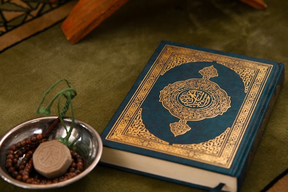 A Quran with an ornate cover is placed on a green fabric. Beside it, a small metallic bowl contains prayer beads and a clay tablet, both typically used in Islamic rituals.