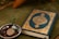 A Quran with an ornate cover is placed on a green fabric. Beside it, a small metallic bowl contains prayer beads and a clay tablet, both typically used in Islamic rituals.