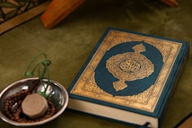 A Quran with an ornate cover is placed on a green fabric. Beside it, a small metallic bowl contains prayer beads and a clay tablet, both typically used in Islamic rituals.