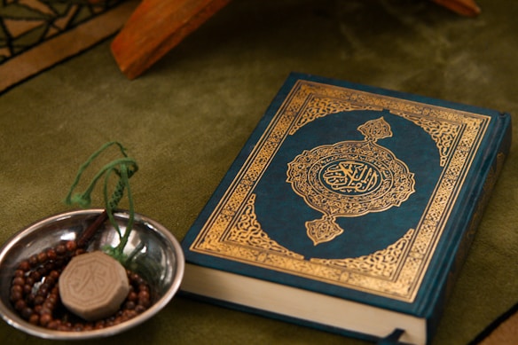 A Quran with an ornate cover is placed on a green fabric. Beside it, a small metallic bowl contains prayer beads and a clay tablet, both typically used in Islamic rituals.