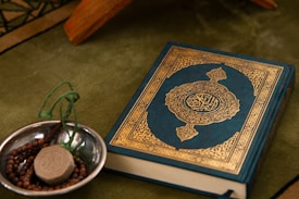 A Quran with an ornate cover is placed on a green fabric. Beside it, a small metallic bowl contains prayer beads and a clay tablet, both typically used in Islamic rituals.
