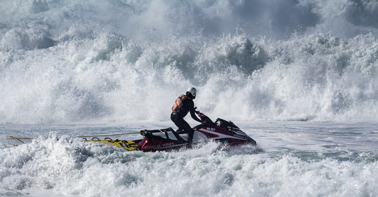A person wearing protective gear and a helmet is riding a jet ski through turbulent, foamy waves. The powerful motion of the water surrounds the rider, creating a dramatic scene of adventure and challenge.