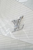 A metallic, reflective logo is mounted on a white, tiled wall. The logo is three-dimensional with a shiny surface, casting subtle reflections onto the surface behind it. The tiles are arranged in a regular pattern, creating a clean and modern aesthetic.