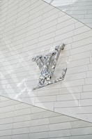 A metallic, reflective logo is mounted on a white, tiled wall. The logo is three-dimensional with a shiny surface, casting subtle reflections onto the surface behind it. The tiles are arranged in a regular pattern, creating a clean and modern aesthetic.
