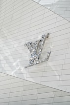 A metallic, reflective logo is mounted on a white, tiled wall. The logo is three-dimensional with a shiny surface, casting subtle reflections onto the surface behind it. The tiles are arranged in a regular pattern, creating a clean and modern aesthetic.