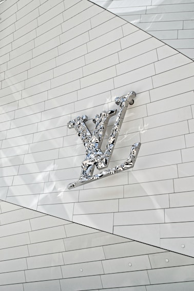 A metallic, reflective logo is mounted on a white, tiled wall. The logo is three-dimensional with a shiny surface, casting subtle reflections onto the surface behind it. The tiles are arranged in a regular pattern, creating a clean and modern aesthetic.