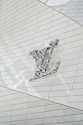 A metallic, reflective logo is mounted on a white, tiled wall. The logo is three-dimensional with a shiny surface, casting subtle reflections onto the surface behind it. The tiles are arranged in a regular pattern, creating a clean and modern aesthetic.