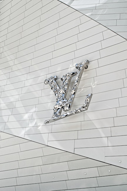 A metallic, reflective logo is mounted on a white, tiled wall. The logo is three-dimensional with a shiny surface, casting subtle reflections onto the surface behind it. The tiles are arranged in a regular pattern, creating a clean and modern aesthetic.