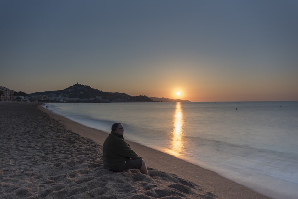 Mann in schwarzem Kapuzenpullover sitzt bei Sonnenuntergang auf braunem Sand in der Nähe von Wasser