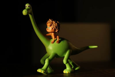 A small figurine depicts a green dinosaur with a long neck and tail. Seated on its back is a small, animated character with shaggy brown hair. The scene is illuminated with a warm light creating a shadow effect.
