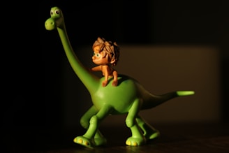 A small figurine depicts a green dinosaur with a long neck and tail. Seated on its back is a small, animated character with shaggy brown hair. The scene is illuminated with a warm light creating a shadow effect.