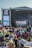 A large outdoor music festival with a crowd seated on the grass, facing a stage with a large screen displaying a musician. The stage is set up with banners that read 'Pori Jazz 2019' and 'International Jazz Festival Every Year in July'. The audience is a mix of standing and seated individuals, many of whom are using picnic chairs. The atmosphere is casual and relaxed.