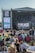 A large outdoor music festival with a crowd seated on the grass, facing a stage with a large screen displaying a musician. The stage is set up with banners that read 'Pori Jazz 2019' and 'International Jazz Festival Every Year in July'. The audience is a mix of standing and seated individuals, many of whom are using picnic chairs. The atmosphere is casual and relaxed.