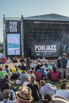 A large outdoor music festival with a crowd seated on the grass, facing a stage with a large screen displaying a musician. The stage is set up with banners that read 'Pori Jazz 2019' and 'International Jazz Festival Every Year in July'. The audience is a mix of standing and seated individuals, many of whom are using picnic chairs. The atmosphere is casual and relaxed.