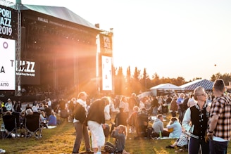 A large outdoor music festival with a crowd gathered on a grassy field during sunset. People are sitting or standing, some on blankets or folding chairs, watching a concert on a large stage with banners reading 'Pori Jazz 2019'. The warm golden light from the sun creates a relaxed and atmospheric ambiance.
