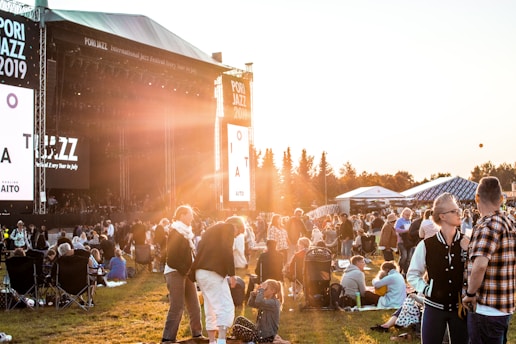 A large outdoor music festival with a crowd gathered on a grassy field during sunset. People are sitting or standing, some on blankets or folding chairs, watching a concert on a large stage with banners reading 'Pori Jazz 2019'. The warm golden light from the sun creates a relaxed and atmospheric ambiance.