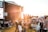 A large outdoor music festival with a crowd gathered on a grassy field during sunset. People are sitting or standing, some on blankets or folding chairs, watching a concert on a large stage with banners reading 'Pori Jazz 2019'. The warm golden light from the sun creates a relaxed and atmospheric ambiance.