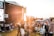 A large outdoor music festival with a crowd gathered on a grassy field during sunset. People are sitting or standing, some on blankets or folding chairs, watching a concert on a large stage with banners reading 'Pori Jazz 2019'. The warm golden light from the sun creates a relaxed and atmospheric ambiance.