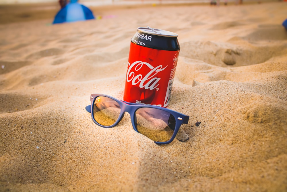 coca cola can beside black framed eyeglasses on brown sand