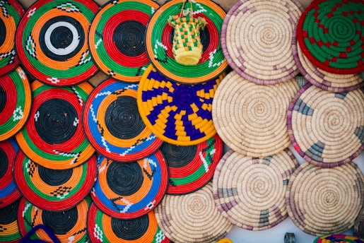 Various circular woven baskets are arranged in a vibrant display. The baskets showcase intricate patterns and a wide array of colors, including red, green, orange, blue, yellow, and black. Some of the baskets feature simple, natural tones with subtle accents.