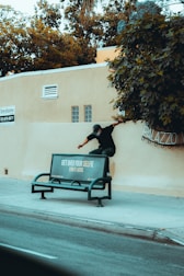 A person is climbing over a beige wall next to a public bench. The bench has an advertisement that reads 'GET OVER YOUR SELFIE SEND IT LATER.' The wall is adjacent to a sidewalk and a street. There are trees and greenery above the wall.