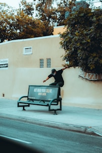 A person is climbing over a beige wall next to a public bench. The bench has an advertisement that reads 'GET OVER YOUR SELFIE SEND IT LATER.' The wall is adjacent to a sidewalk and a street. There are trees and greenery above the wall.