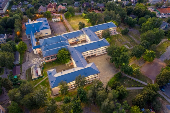 An aerial view of a large, L-shaped building complex surrounded by trees and pathways. The rooftops are blue, and the building is illuminated by outdoor lighting. The surrounding area includes other smaller buildings, open spaces, and residential houses.